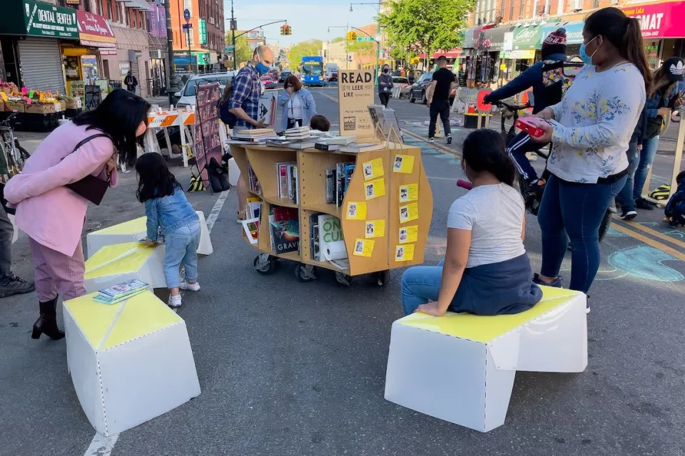 Children sit on temporary street furniture next to book cart at pop-up reading event at open streets event in Sunset Park in New York City.